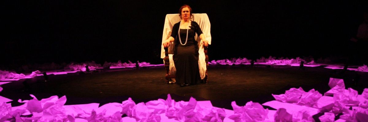 Hedda is sitting in a chair in the middle of an open circle surrounded by crunched paper lit in purple.