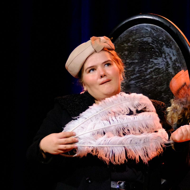 A lady wearing an elegant hat holding a white feather with her back to a round mirror.