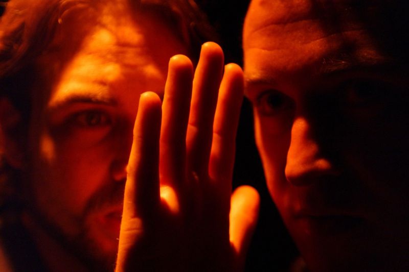 Peer Gynt and the Button-Moulder are looking at a close-up of a hand in an amber light that highlights the hand suggesting a disconcerting importance.