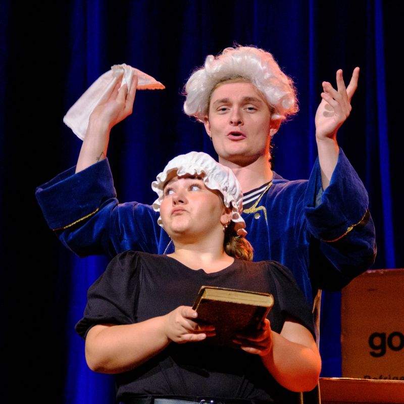 An aristocrat wearing a white baroque wig and his maid wearing a simple cap and holding a book interact.