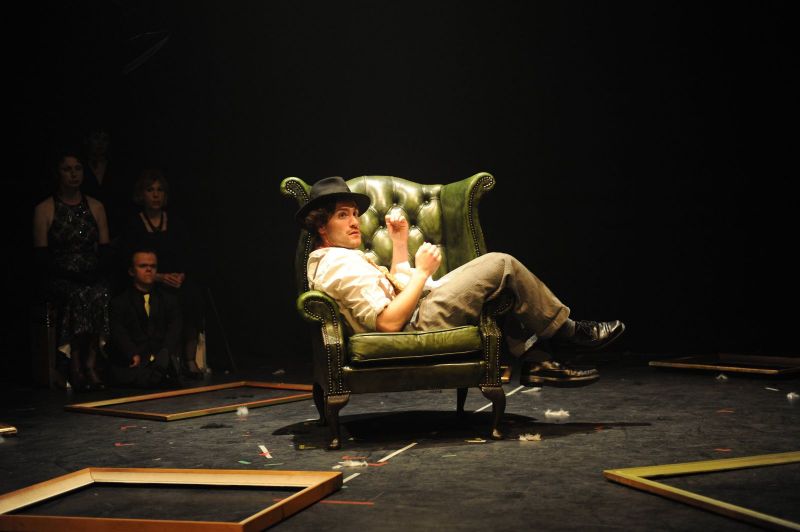 Peer Gynt is in profile. He is sitting in a green scholar armchair with his feet across the armrest. Picture frames are scattered on floor of the entire stage.