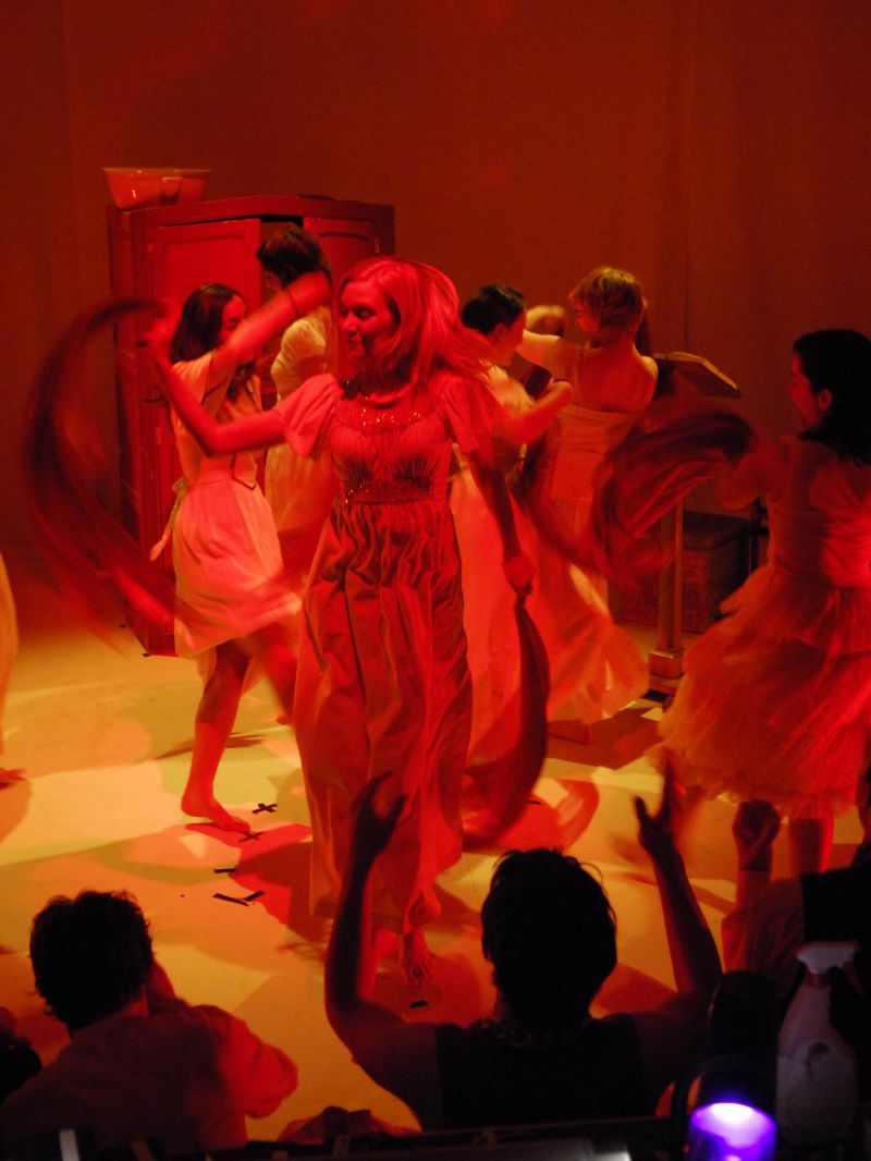 Anitra and a harem of girls are perfoming a dance with their veils barefoot. The lighting is a mix of amber and red.