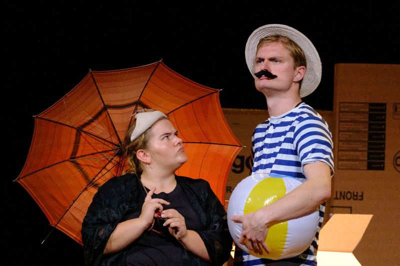 A Victorian lady wearing a hat and an umbrella and a gentleman with a moustache wearing a straw hat and a striped swimming costume holding a ball are posing for the camera. The lady looks at the gentleman. Her facial expression is annoyed.