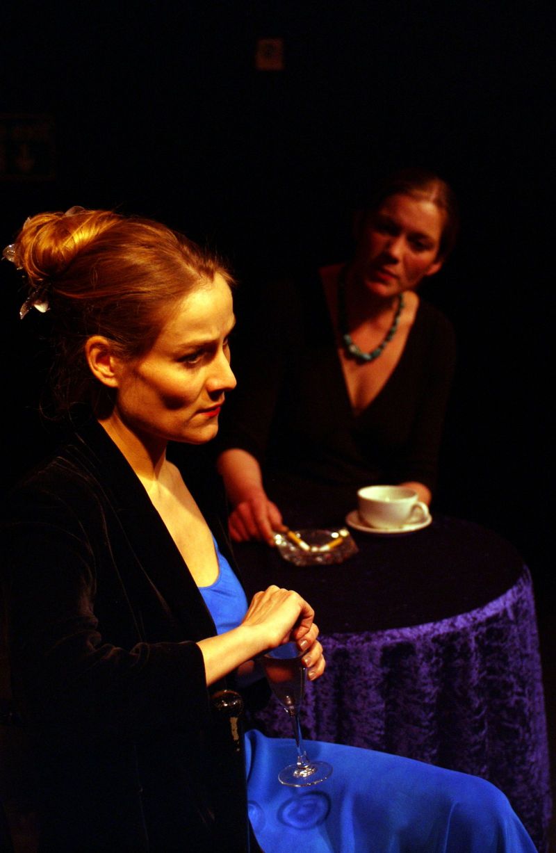 Two women (Amelia and Madam) are discussing across a round table with a dark blue velvet tablecloth, a single coffee cup and an ashtray.