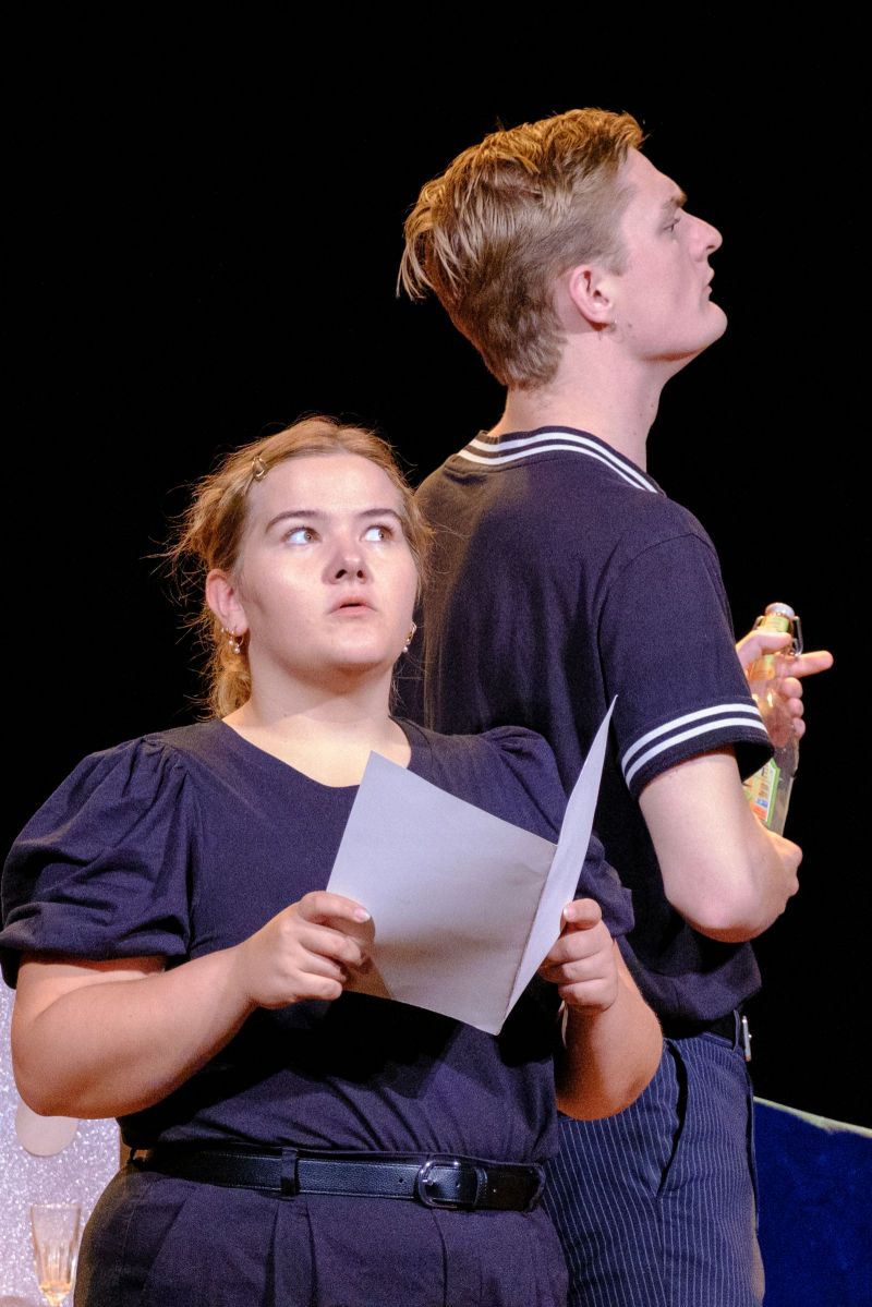 A young woman is facing us holding a pice of paper. Her facial expression is serious and contemplative. Next to her, in profile, a young man holding a bottle.