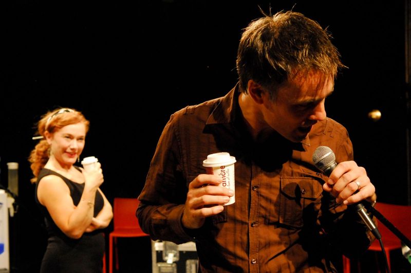 Brack is having fun speaking into a microphone in the recording studio while Hedda is amused and watching in the background with a takeaway coffee.