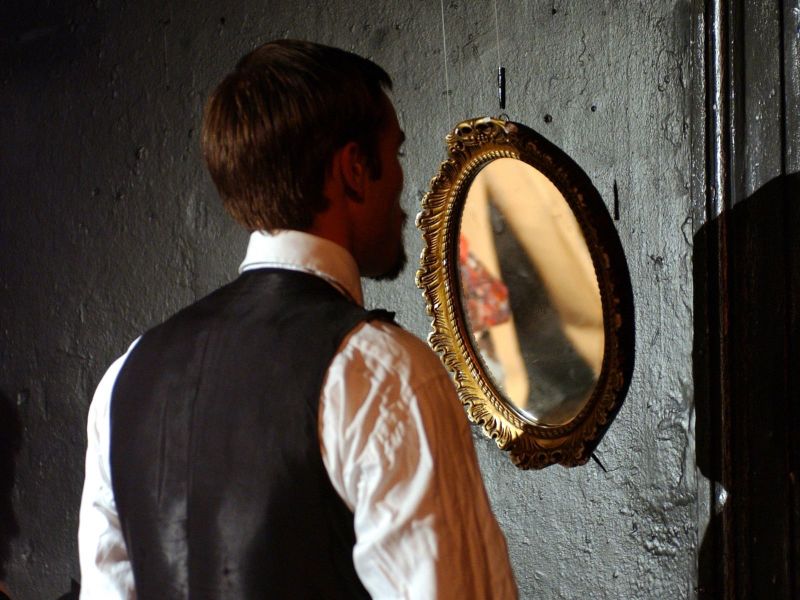 Bernick, with his back towards us, is looking at himself in round mirror with a golden frame.
