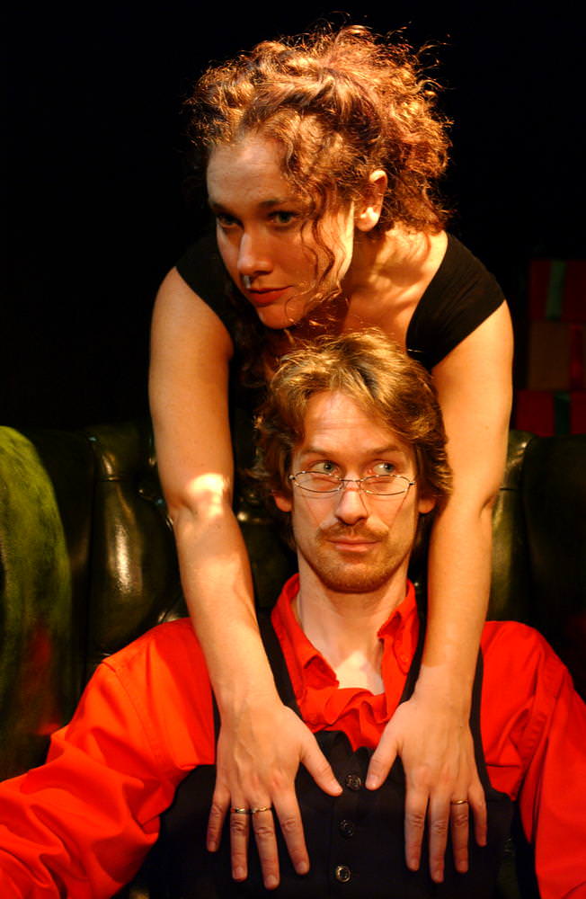 Helmer is sitting in a leather arm chair wearing reading glasses. Nora is standing behind him stretching her arms onto his chest.