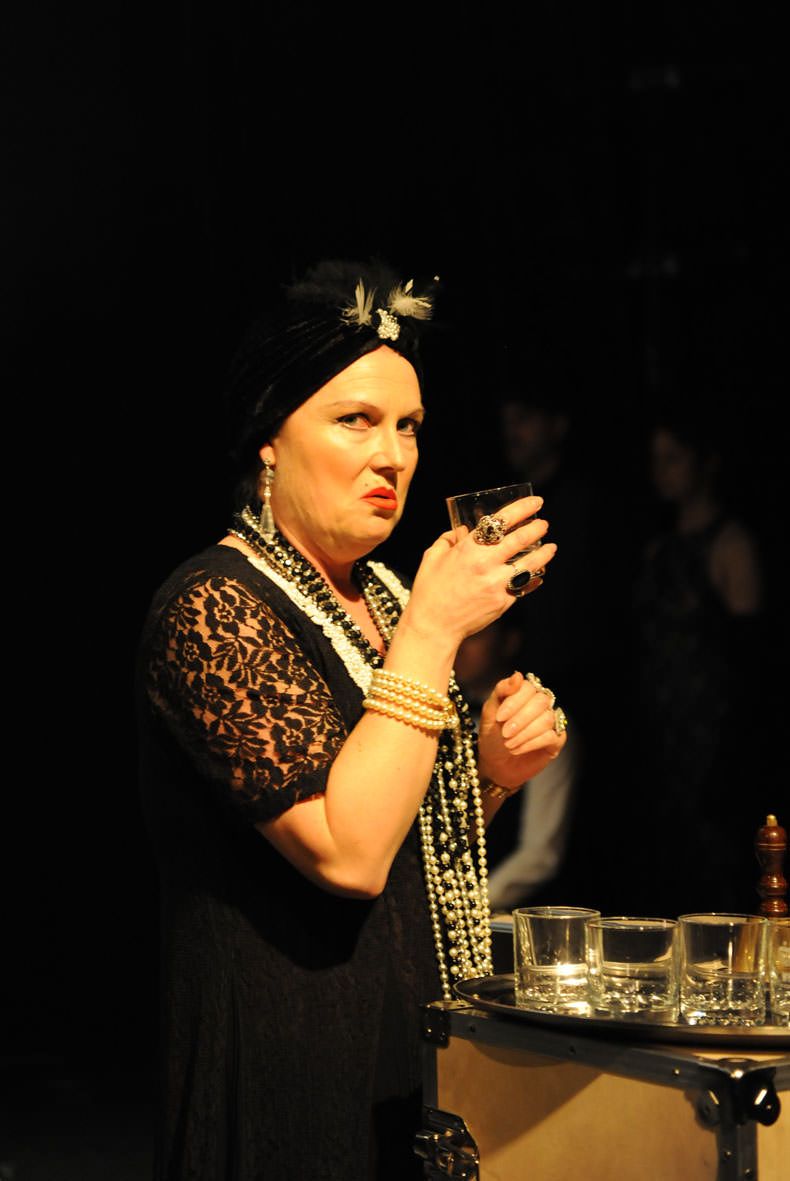 Peer Gynt&#039;s mother, Åse, is having a drink on her own. She is dressed in old cocktail dress with her entire collection of jewellery around her neck.